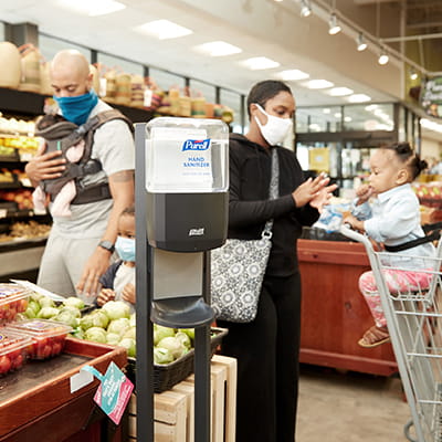 Family wearing face masks shops at grocery store, uses PURELL hand sanitizer dispenser