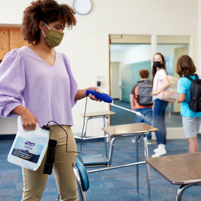 Teacher sprays PURELL surface disinfectant on desks with students present