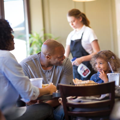 Family seated at a table dining at casual restaurant with an employee behind them spraying another table with PURELL Foodservice Surface Sanitizer