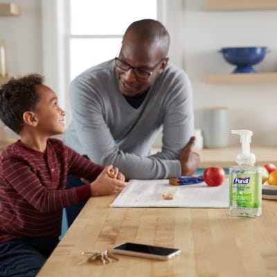 Young child uses PURELL® Advanced Hand Sanitizer Naturals Foam before snack time in the kitchen, while dad talks with him